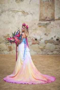 a woman with flowers in her hair is standing near a stone wall and holding a bouquet