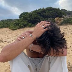 a man sitting on top of a sandy beach holding his hands to his head with trees in the background