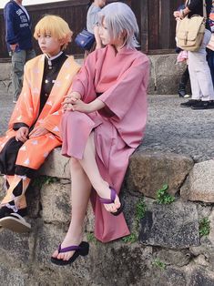 two people in kimonos sitting next to each other on some rocks with others standing around