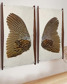 two wall hangings decorated with gold and brown feathers on white paper next to a wooden shelf