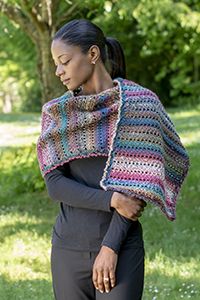 a woman wearing a multicolored crocheted shawl in the grass with her hands on her hips