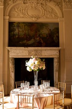 a dining room table set with flowers and wine glasses in front of an ornate fireplace