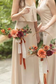 two bridesmaids in long dresses holding flowers and hoop wreaths with feathers on them