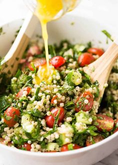 a white bowl filled with couscous, tomatoes and cucumbers being drizzled with dressing