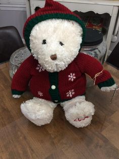 a white teddy bear wearing a red sweater and green hat sitting on top of a wooden table