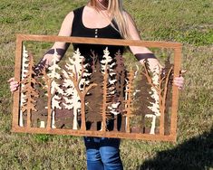 a woman holding up a cut out of wood with trees in the woods on it