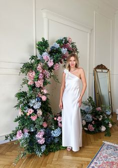 a woman in a white dress standing next to a floral arch with pink and blue flowers