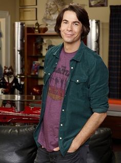 the young man is posing for a photo in his living room, wearing a green shirt and black pants