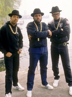 three men in black hats and chains standing next to each other near the water with their arms crossed