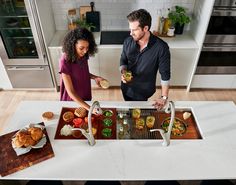 a man and woman preparing food on a counter top in a kitchen with stainless steel appliances