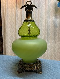 a green glass vase sitting on top of a blue tablecloth covered table next to a white curtain