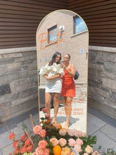 two women standing in front of a mirror with flowers on the ground next to it