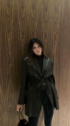 a woman standing in front of a wooden wall holding a handbag and posing for the camera