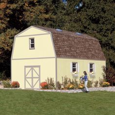 a man standing in front of a yellow barn with flowers on the lawn and trees behind him