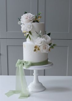 a wedding cake with white flowers and green ribbons on a table in front of a gray wall