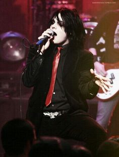 a man with black hair and red tie singing into a microphone