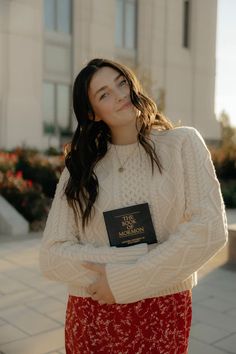 a woman standing in front of a building with her arms crossed, wearing a white sweater and red skirt