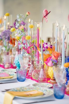 the table is set with many colorful vases and flowers in glass bottles, candles, and napkins