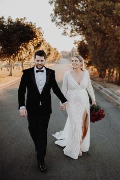 a bride and groom walking down the road holding hands in their tuxedo outfits