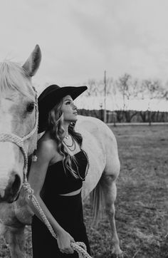 a woman in a black dress and hat standing next to a white horse with a rope