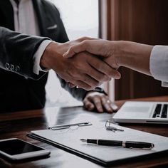 two people shaking hands over a desk with a laptop and notebook on it, while another person holds the hand of another person's hand