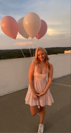 a woman in a pink dress is holding three balloons and posing for the camera with her hands behind her back