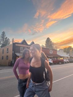 two women standing on the side of a road with their arms around each other as the sun sets behind them