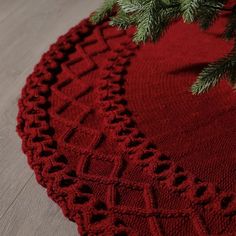 a close up of a red knitted placemat on a wooden floor next to a pine tree