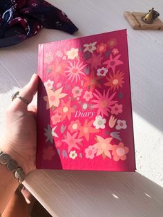 a person holding up a pink card with flowers on it and the word diary written in large letters