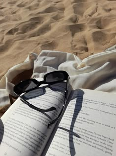 an open book with sunglasses on top of it sitting in the sand at the beach