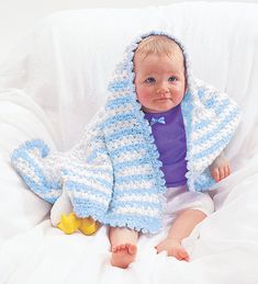 a baby wearing a blue and white crochet jacket sitting on a bed with a rubber duck