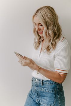 a woman in white shirt and jeans holding a cell phone with her right hand on her hip