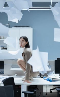 a woman kneeling down on top of a desk in an office with papers flying around her