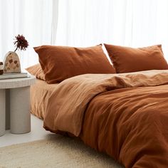 a bed with orange sheets and pillows next to a white table in front of a window