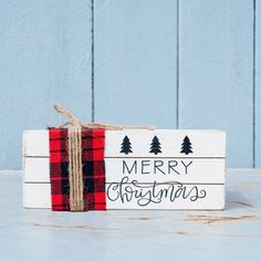 a merry christmas sign with pine trees and plaid ribbon tied around the edges on a blue background