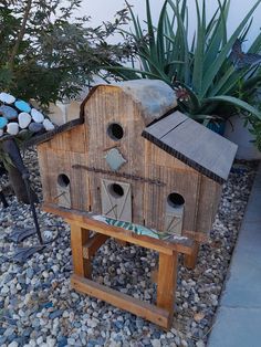 a wooden bird house sitting on top of a gravel covered ground next to plants and rocks