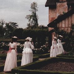 four women in white dresses and hats are standing on the lawn near a building with hedges