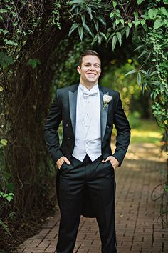 a man in a tuxedo poses for a photo under an archway with greenery