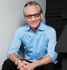 a man sitting on top of a chair wearing glasses and a blue shirt with his hands in his pockets