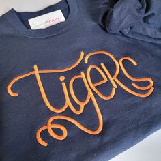 Calling all Auburn Tigers fans! This navy blue sweatshirt with bold orange 'Tigers' embroidery is your go-to choice for game day or everyday style. Stay cozy and show your allegiance to Auburn University. Available in unisex sizes S-3XL. War Eagle! Personalized Slippers, Navy Blue Sweatshirt, Tiger Logo, Baby Beanie Hats, College Shirts, Baby Bloomers, Auburn University, Auburn Tigers, Embroidered Clothes