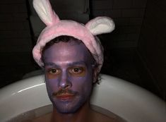 a man with purple paint on his face in a bathtub wearing a bunny hat