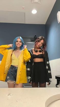 two young women standing in front of a mirror wearing yellow jackets and black shorts, one with blue hair