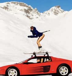 a woman is standing on top of a red sports car in front of snow covered mountains