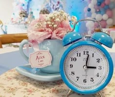 a blue alarm clock sitting on top of a table next to a teacup filled with flowers