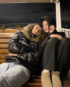 two people sitting on a bench with their arms around each other, both wearing winter coats