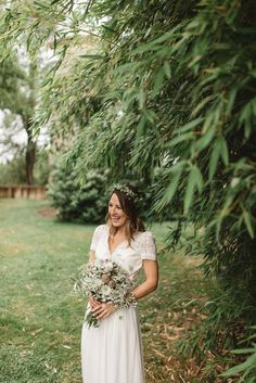 a woman in a white dress holding a bouquet standing next to some trees and grass