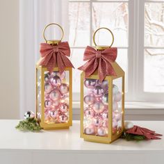 two christmas lanterns with bows and baubles in them on a table next to a window