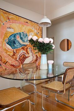 a glass dining table with chairs and a vase on it in front of a painting