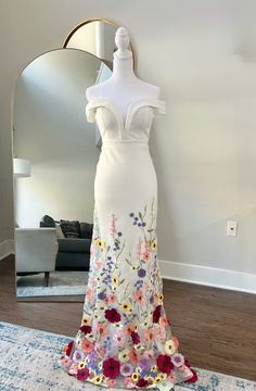 a white dress with flowers on it is displayed in front of a mirror and rug