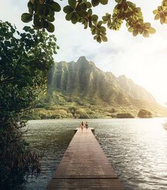 two people standing on a dock in front of the water with mountains in the background